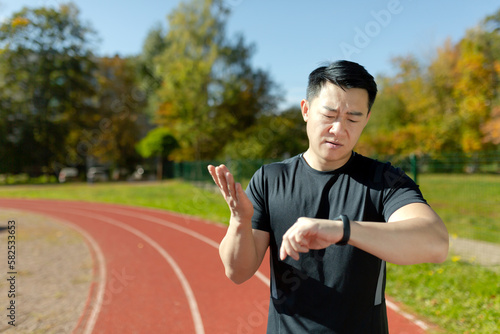 Portrait of a young Asian male athlete  a runner standing on a treadmill in a stadium and checking the time on a smart watch. Dissatisfied with the result  he spreads his hands in frustration.