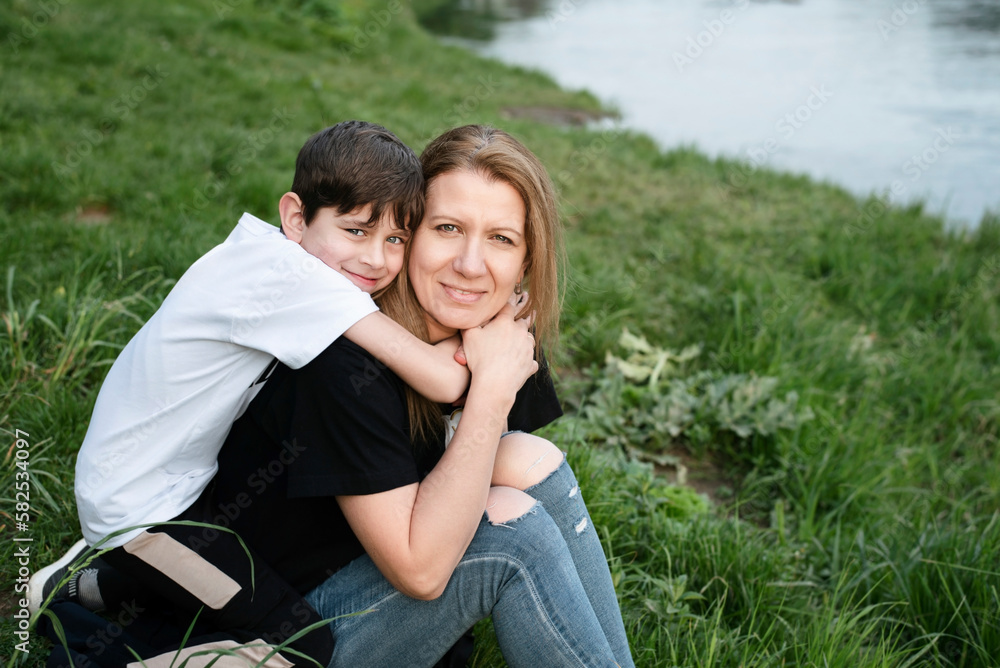 Fototapeta premium Mother and little son spend time together on the river bank in spring, the boy is not yet a teenager and still loves his parents very much, often hugs her, says how much he loves