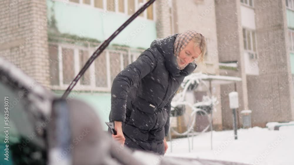 Portrait of young woman removing snow from roof and windows of car with scraper and counter. Heavy snowfall and cyclone. Retarded motion. Snow falls, against backdrop of city. Portrait of young woman removing snow from roof and windows of car with scraper and counter. Heavy snowfall and cyclone. Retarded motion. Snow falls, against backdrop of city.