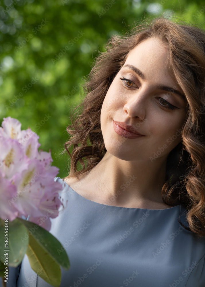 A beautiful young girl with dark hair in a gray dress looks at a rhododendron flower. Close-up. Tenderness, thoughtfulness