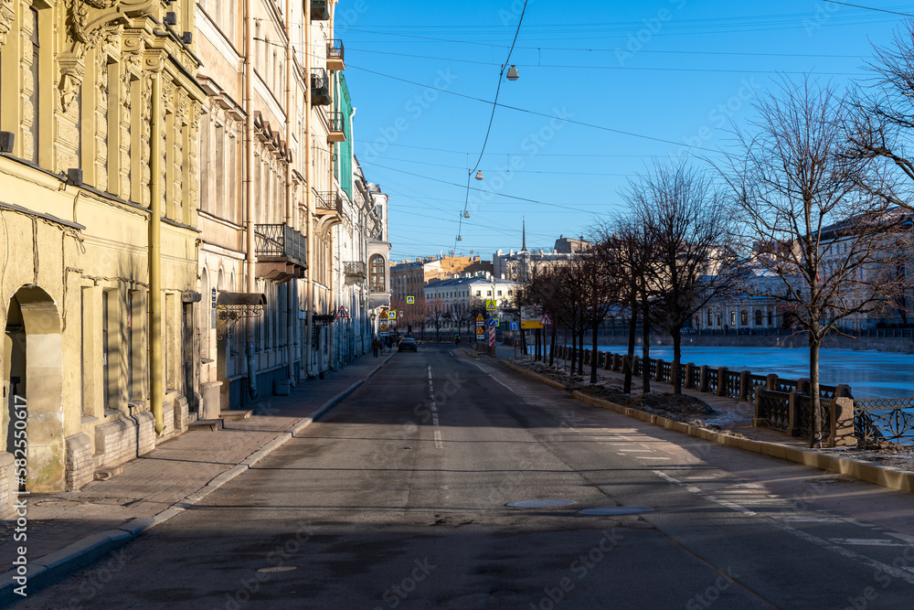 Emty street with bare trees on Fontanka river embankment in sunny winter morning in Saint Petersburg city, Russia. River surface covered with ice. Soft focus. Early morning traffic theme.