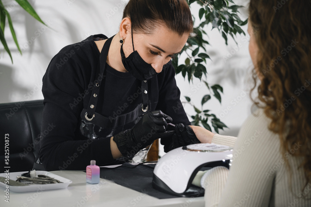 Manicure salon. Professional manicurist wearing protective mask doing procedure for woman in modern beauty salon. Beauty industry concept. Lamp for polish Manicure salon. Professional manicurist wearing protective mask doing procedure for woman in modern beauty salon. Beauty industry concept. Lamp for polish