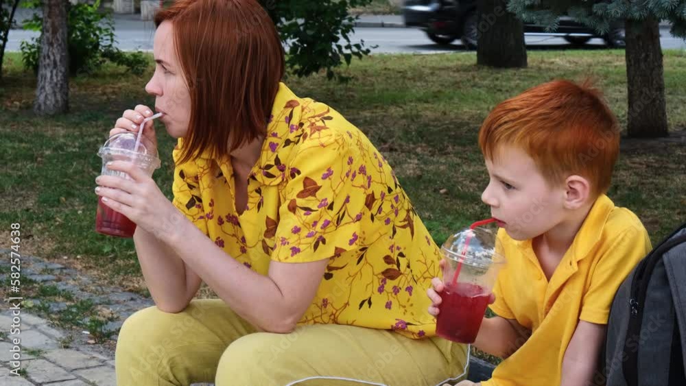Mother and young son are drinking a cocktail in the park. Middle-aged red-haired woman and red-haired young boy are resting and talking. Mother and young son are drinking a cocktail in the park. Middle-aged red-haired woman and red-haired young boy are resting and talking.