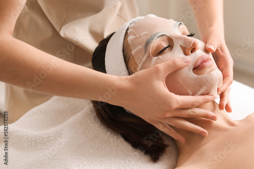 Fotografía Cosmetologist applying sheet mask onto young woman's face in salon, closeup