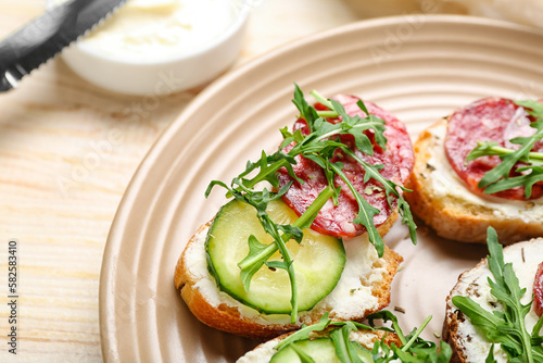 Plate of tasty sandwiches with cream cheese, cucumber, arugula and salami on wooden table, closeup