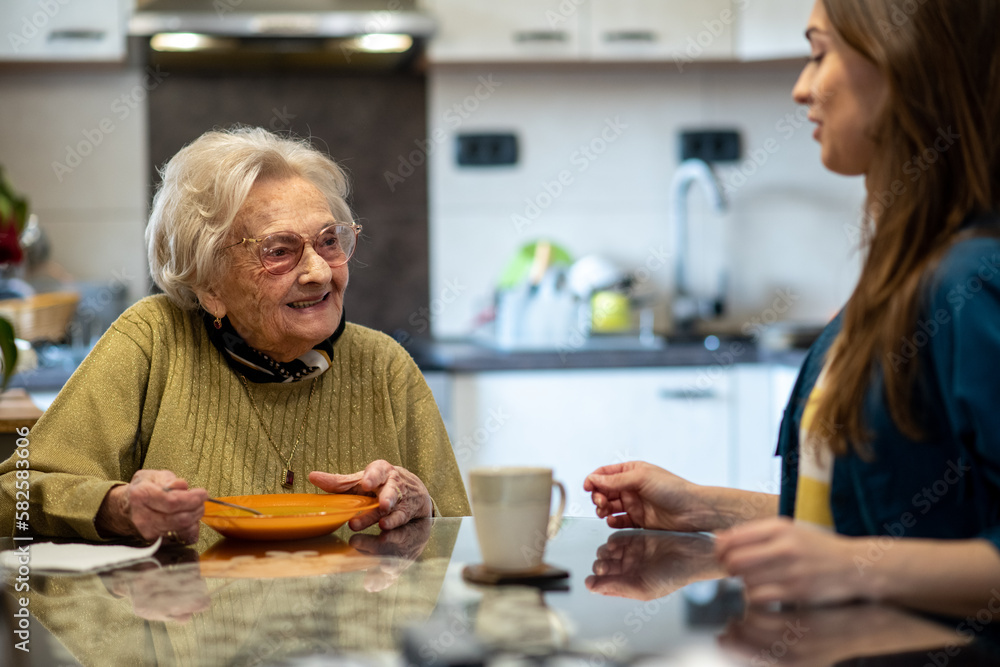 Senior woman eating soup at a dining table while caretaker nurse sits next to her at a nursing home.