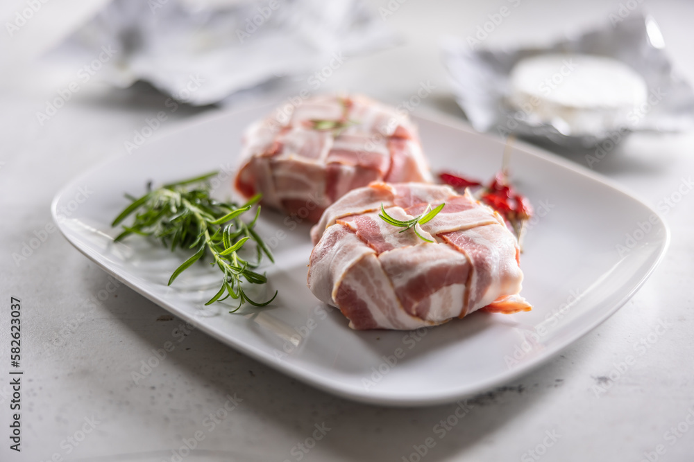 Camembert or brie cheeses wrapped in bacon with fresh rosemary and chilli on a white plate