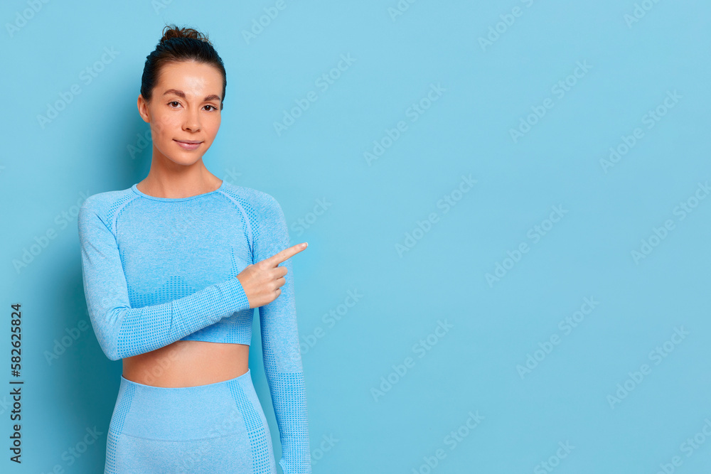 Cute brunette in a blue yoga costume stands against a blue background and points her finger at the open space for your ad, copy space, trade concept, high quality photo Cute brunette in a blue yoga costume stands against a blue background and points her finger at the open space for your ad, copy space, trade concept, high quality photo