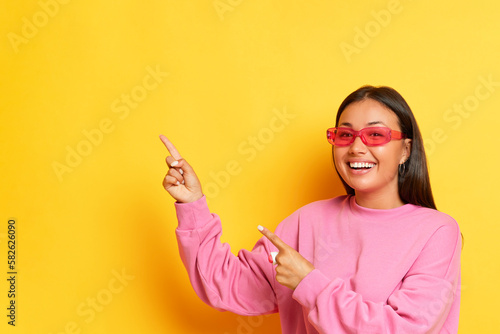 Nice smiling female model in pink sunglasses and pink sweatshirt posing on yellow backdrop  pointing her fingers at copy space aside  high quality photo.