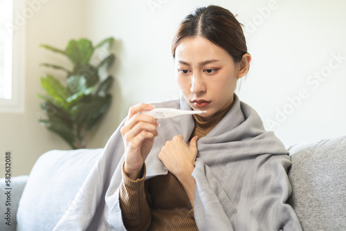 Sick, influenza asian young woman, girl headache have fever, hand touching forehead, holding thermometer for check measure body temperature, illness while sitting rest on sofa at home. Health care.