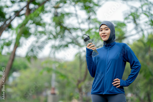 Asian cute muslim woman wearing sportswear and a blue hijab doing exercise and being tired and drinking water outdoors at the park in the morning with a fresh feeling.