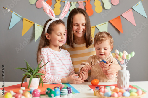 Image of joyful cheerful woman hugging daughters while sitting at table, preparing for Easter, little child dressed in bunny ears and mom looking how infant baby trying to paint eggs..