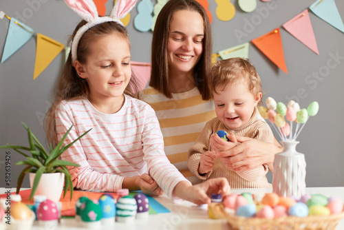 Photo of delighted positive female with brown hair and her children painting eggs, preparing for Easter, spending time together, celebrating spring holiday.