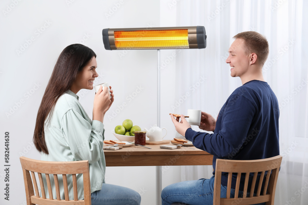 Couple spending time in room with modern electric infrared heater, focus on appliance Couple spending time in room with modern electric infrared heater, focus on appliance