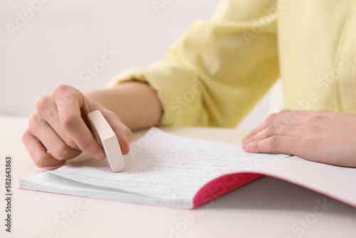 Girl erasing mistake in her notebook at white desk indoors, closeup