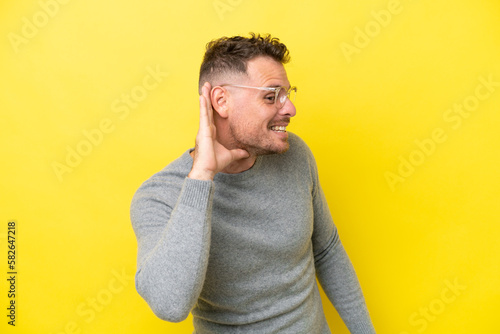 Young caucasian handsome man isolated on yellow background listening to something by putting hand on the ear