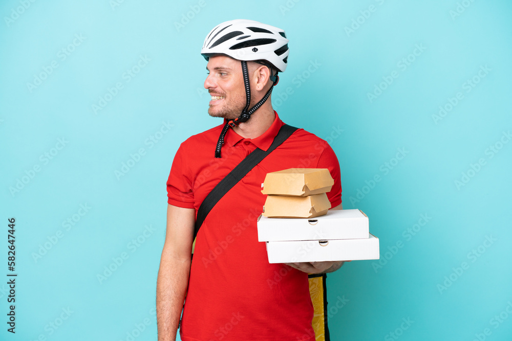 Young caucasian man with thermal backpack and holding fast food isolated on blue background looking side Young caucasian man with thermal backpack and holding fast food isolated on blue background looking side