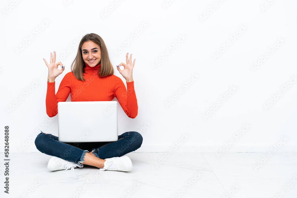 Young caucasian woman sitting on the floor with a laptop showing an ok sign with fingers Young caucasian woman sitting on the floor with a laptop showing an ok sign with fingers