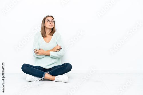 Young caucasian woman sitting on the floor isolated on white background making doubts gesture while lifting the shoulders