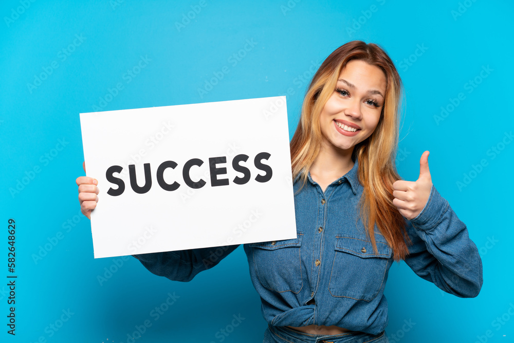 Teenager girl over isolated blue background holding a placard with text SUCCESS with thumb up Teenager girl over isolated blue background holding a placard with text SUCCESS with thumb up