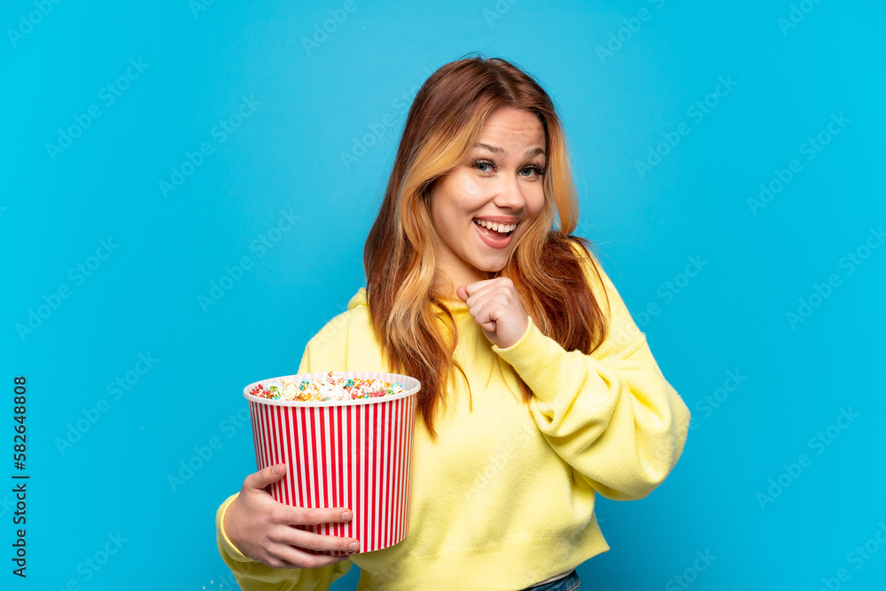 Teenager girl holding popcorns over isolated blue background celebrating a victory Teenager girl holding popcorns over isolated blue background celebrating a victory