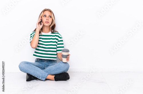 Young Russian girl sitting on the floor isolated on white background holding coffee to take away and a mobile