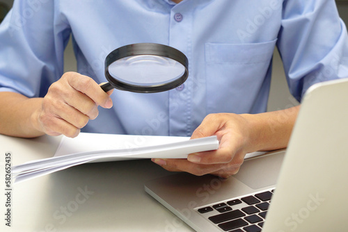 Close up hand of businessperson looking at documents through magnifying glass and laptop in front. Concept of business and data validation.