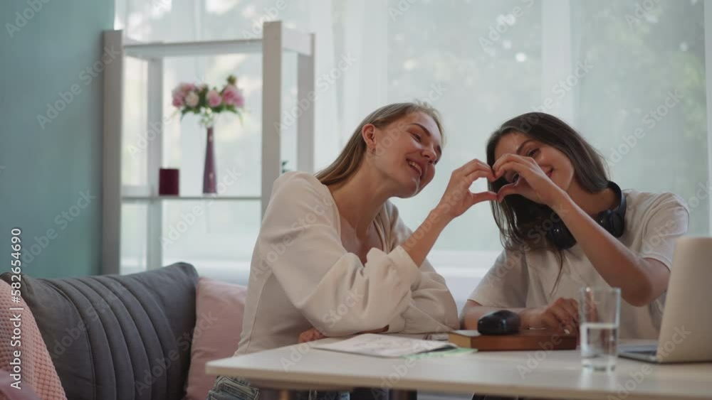 Happy lesbian women make shape of heart with fingers smiling and looking in camera. LGBT couple shows love sitting at table near laptop closeup Happy lesbian women make shape of heart with fingers smiling and looking in camera. LGBT couple shows love sitting at table near laptop closeup