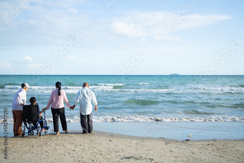 elderly asian woman sitting on wheelchair enjoying together time with friends walking on summer beach