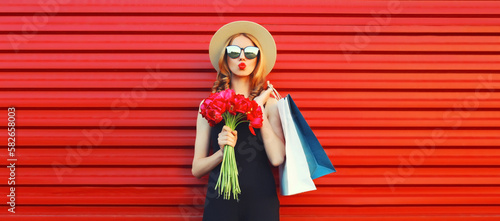 Portrait of beautiful happy young woman with shopping bags and bouquet of flowers wearing summer straw round hat on red background