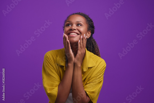 Young happy excited African American woman touching chin and leaning down delighted with invitation to summer trip from boyfriend or hearing other good news standing posing on purple background