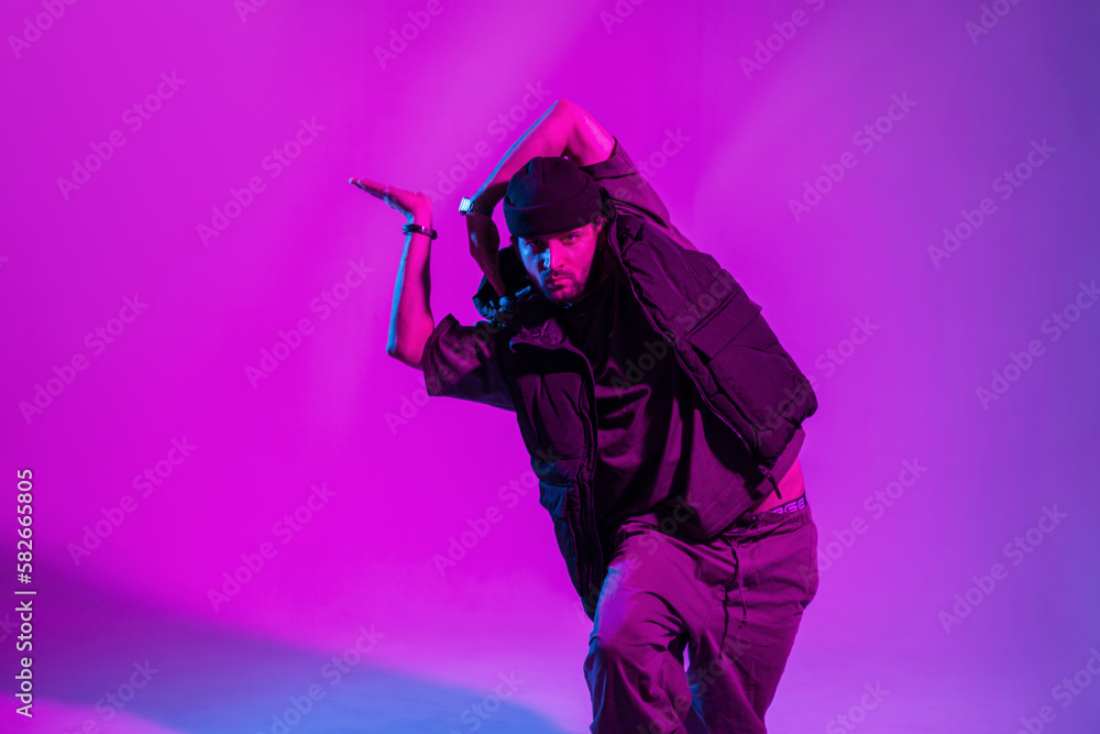 Fashionable professional male dancer in trendy fashion clothes with a hat and vest dancing in a creative color studio with pink neon lights