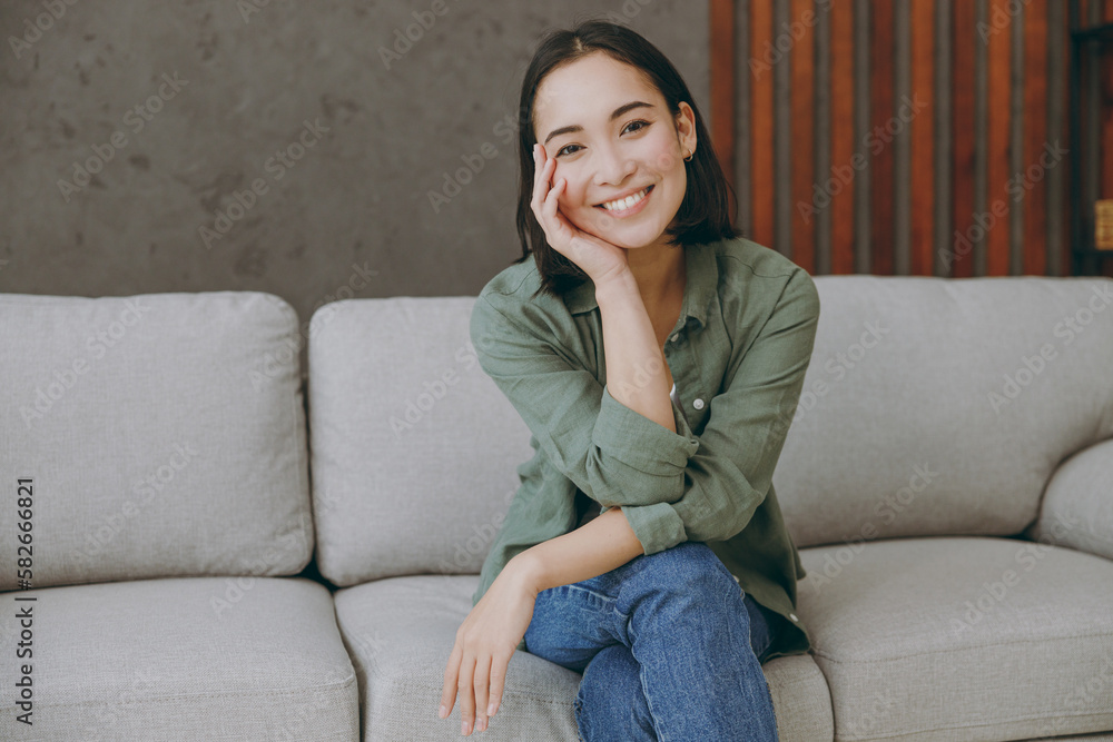 Young smiling fun happy woman of Asian ethnicity wears casual clothes look camera prop up chin sits on grey sofa couch stay at home hotel flat rest relax spend free spare time in living room indoor. Young smiling fun happy woman of Asian ethnicity wears casual clothes look camera prop up chin sits on grey sofa couch stay at home hotel flat rest relax spend free spare time in living room indoor.