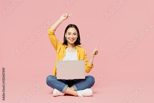 Full body fun young IT woman of Asian ethnicity wear yellow shirt white t-shirt sitting hold use work on laptop pc computer point finger aside on area isolated on plain pastel light pink background.