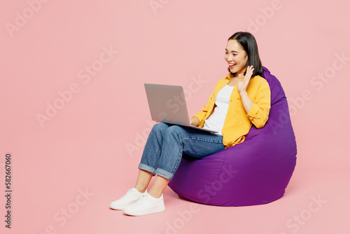 Full body fun young IT woman of Asian ethnicity wear yellow shirt white t-shirt sit in bag chair hold use work on laptop pc computer waving hand isolated on plain pastel light pink background studio.