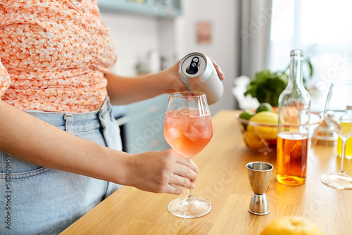 drinks and people concept - close up of woman pouring soda from tin can to wine glass and making orange cocktail at home kitchen