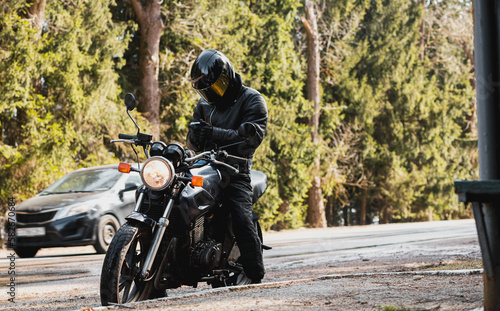 man in a leather motorcycle jacket with a motorcycle custom cafe racer in a helmet on a forest road with a smartphone.