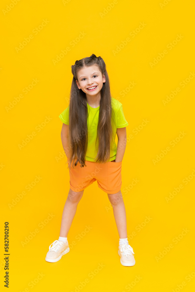 A child in shorts and a T-shirt. Happy childhood. A little girl with long hair enjoys the warmth in summer clothes on a yellow isolated background. A child in shorts and a T-shirt. Happy childhood. A little girl with long hair enjoys the warmth in summer clothes on a yellow isolated background.