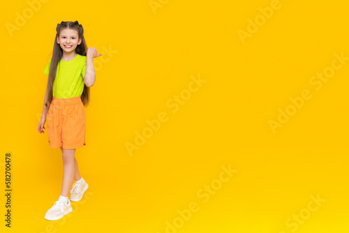 Fototapeta Naklejka Na Ścianę i Meble -  A child in shorts and a T-shirt. Happy childhood. A little girl with long hair enjoys the summer warmth inside and points to your advertisement on a yellow isolated background. Copy space. Banner.