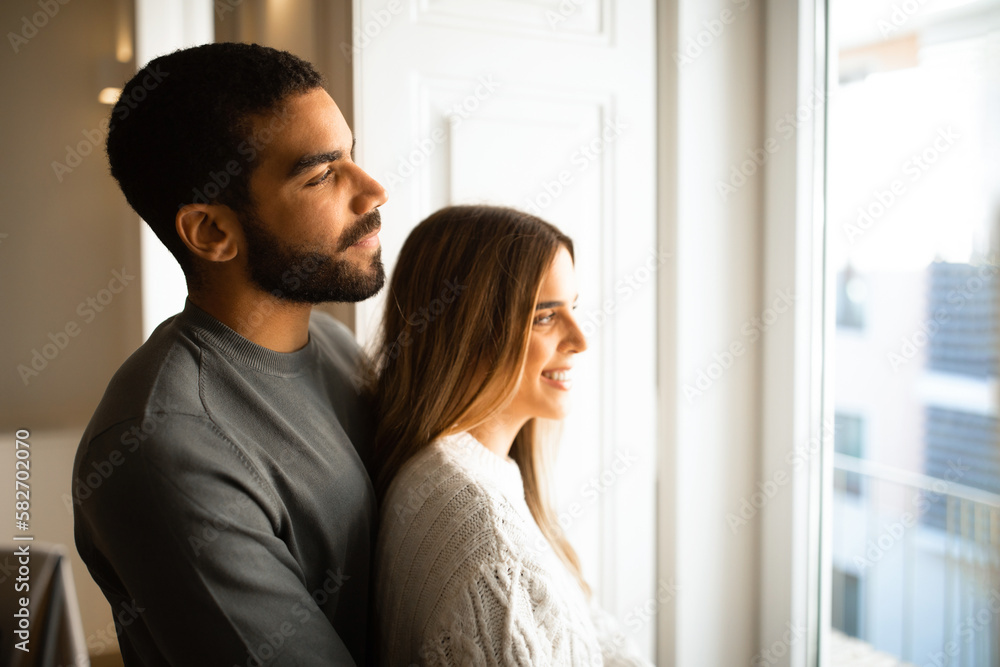 Cheerful international millennial couple hugging, look out the window, enjoy free time together Cheerful international millennial couple hugging, look out the window, enjoy free time together
