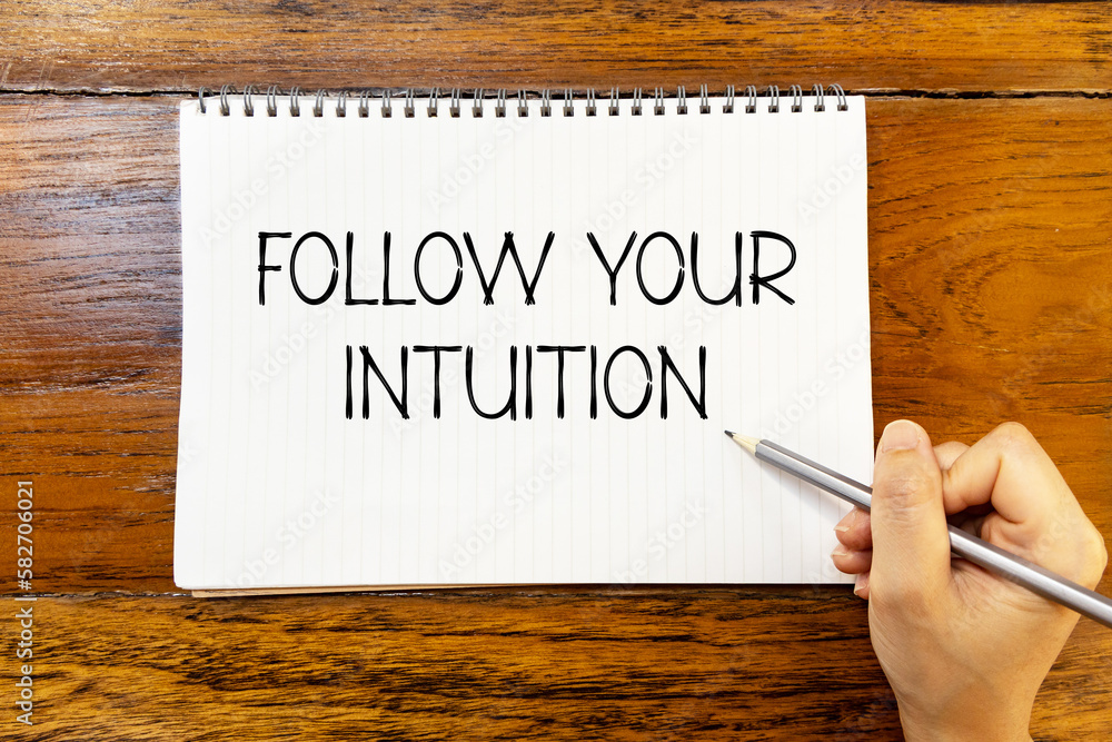 Follow your intuition handwriting text on blank notebook paper on wooden table with hand holding pencil. Business concept about following your intuition. Follow your intuition handwriting text on blank notebook paper on wooden table with hand holding pencil. Business concept about following your intuition.