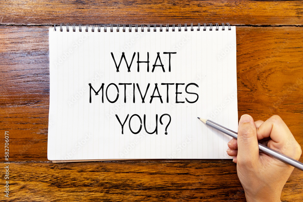 What motivates you handwriting text on blank notebook paper on wooden table with hand holding pencil. Business concept about motivation. What motivates you handwriting text on blank notebook paper on wooden table with hand holding pencil. Business concept about motivation.