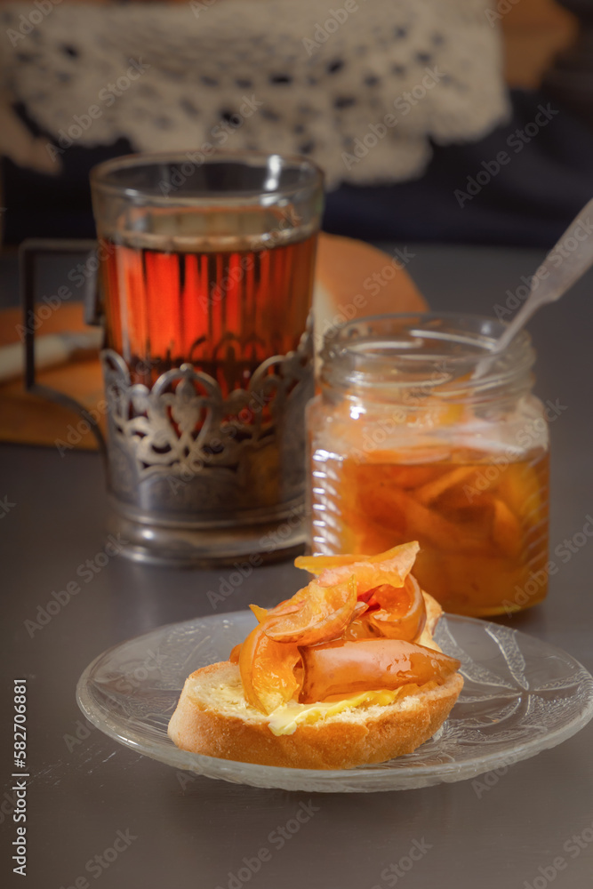 Piece of rustic bread with pear jam, a glass of hot tea in an antique silver cup holder and a jar of jam on a gray wooden table. Minimalism. Village breakfast Piece of rustic bread with pear jam, a glass of hot tea in an antique silver cup holder and a jar of jam on a gray wooden table. Minimalism. Village breakfast