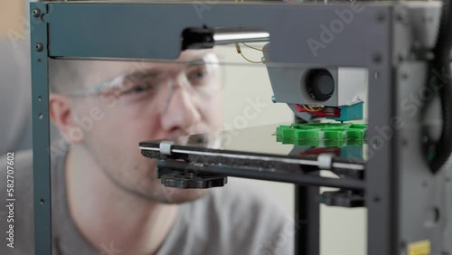 engineer man wearing glasses watching the process of printing the gear detail from green plastic in a 3d printer.