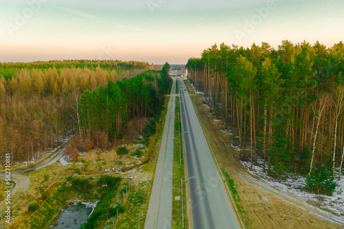 Asfaltowa droga przez sosnowy, wysoki las. Z lewej strony widać latarnie uliczne i chodnik ze ścieżką rowerową. Widok z wysokości, zdjęcie zrobione z użyciem drona.