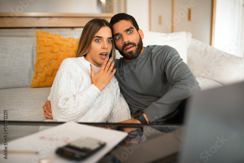 Scared shocked international millennial couple looking at laptop, have video call, pay taxes in living room