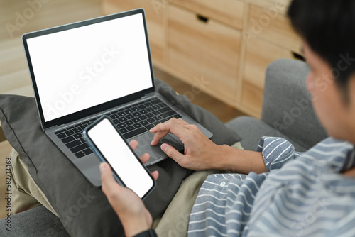Young adult man working online on laptop and using smart phone in living room. Blank screen for advertising text message