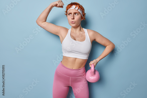 Motivated woman raises arm shows biceps and strength holds weight dressed in cropped top and leggings does exercises in gym looks seriously aside isolated over blue background. Bodybuilding concept
