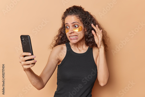 Worried stressful Afro American woman makes selfie photo clenches teeth stares puzzled at front camera dressed in casual black t shirt applies hydrogel golden patches under eyes to reduce puffiness