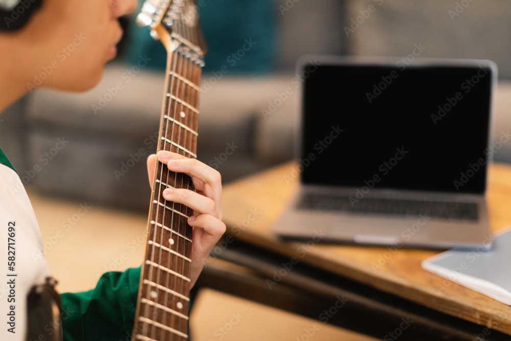 Fototapeta premium Guy Wearing Earphones Playing Electric Guitar Sitting At Laptop Indoors
